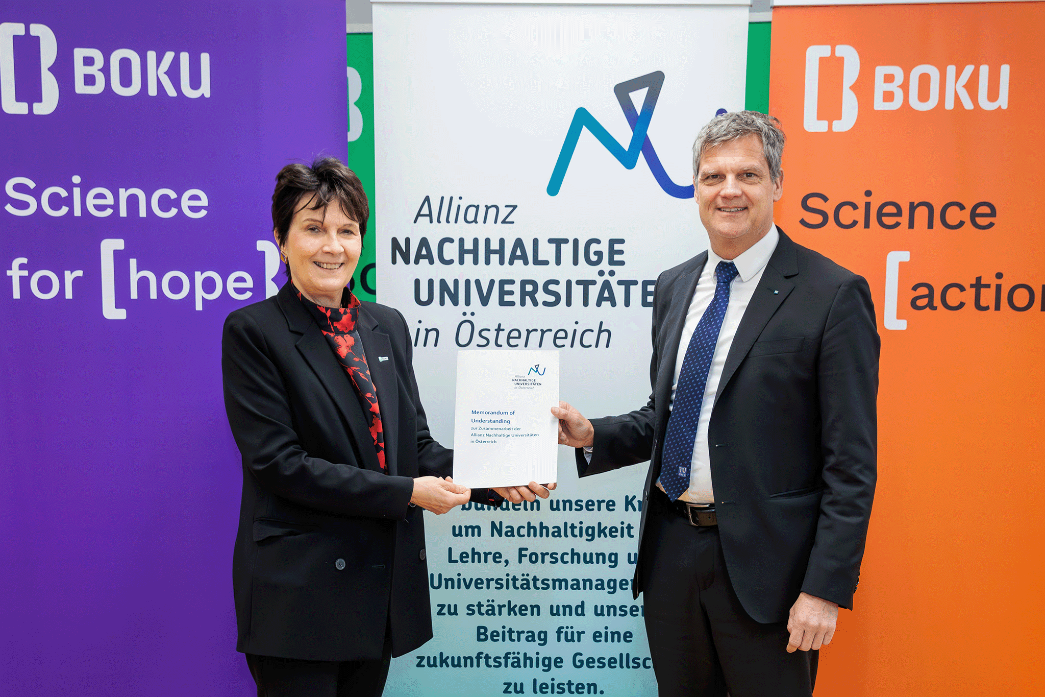 Präsentation des Vertrags. Die Rektorin und der Rektor halten ihn gemeinsam in Händen. Im Hintergrund lila-violettes Boku-Branding.