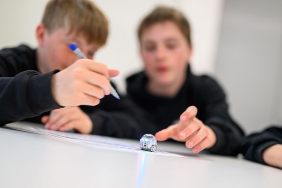 zwei Burschen sitzend bei Tisch, dunkle Oberteile, programmieren kleinen runden Roboter