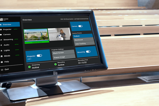 Ein Computermonitor zeigt eine Systemsteuerungsoberfläche mit mehreren blauen Button und Einstellungsoptionen an. Er steht in einem leeren Hörsaal und vermittelt eine technikorientierte Atmosphäre.