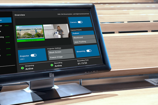Ein Computermonitor zeigt eine Systemsteuerungsoberfläche mit mehreren blauen Button und Einstellungsoptionen an. Er steht in einem leeren Hörsaal und vermittelt eine technikorientierte Atmosphäre.