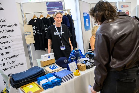 TU Merchandise auf Tisch mit Menschen davor/dahinter