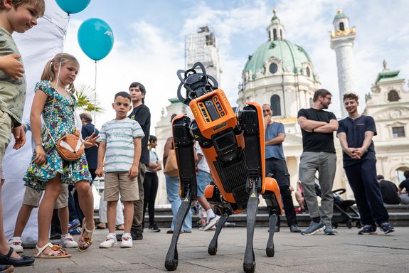 Orangener Roboterhund legt Kopf schief. Danaben emehrere Kinder mit Luftballons
