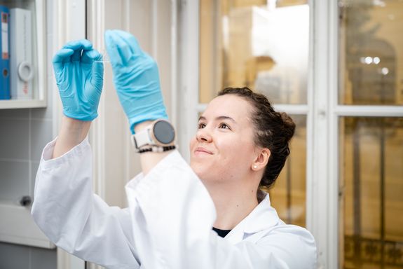 Junge Forscherin in weißem Mantel und türkisen Handschuhen hält eine Laborprobe online und lächelt dabei.
