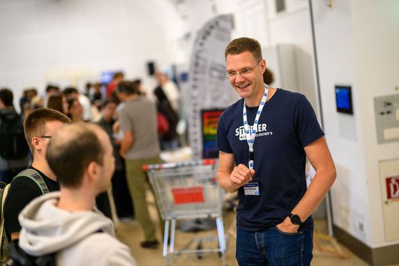 Mann in dunklem Shirt lachend, davor Menschen