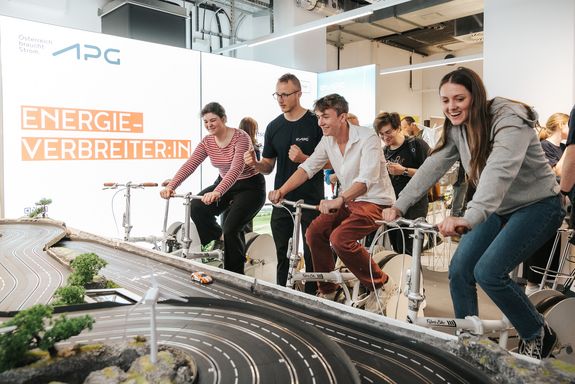 3 junge Menschen sitzen auf Rädern und erzeugen radfahrend Strom. Dadurch werden die Matchboxautos auf der Rennstrecke vor ihnen angetrieben. 