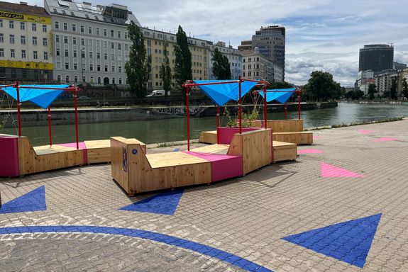 Außenaufnahme Beton mit Bemalung in blau und pink, dahinter Sitzbänke aus Holz